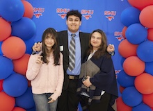 A young woman and a young man stand near a red-and-blue balloon arch making the "pony ears" hand gesture, while an older woman holding a mortarboard stands next to them.