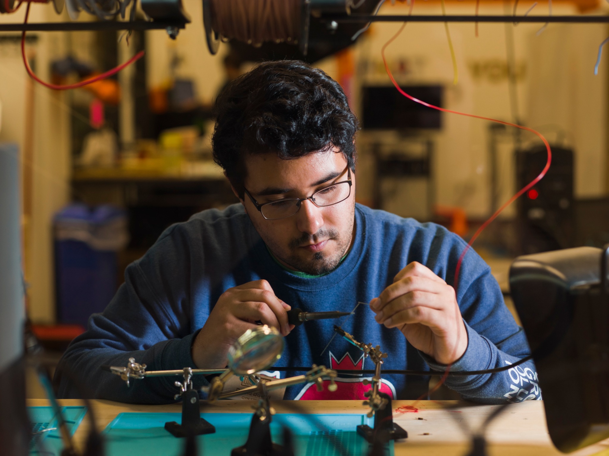 A person with glasses and dark hair is focused on soldering electronic components at a workstation. The person is wearing a blue sweater, and various wires, tools, and a magnifying glass are visible on the table. The background shows a workshop environment with electronic equipment and hanging wires.