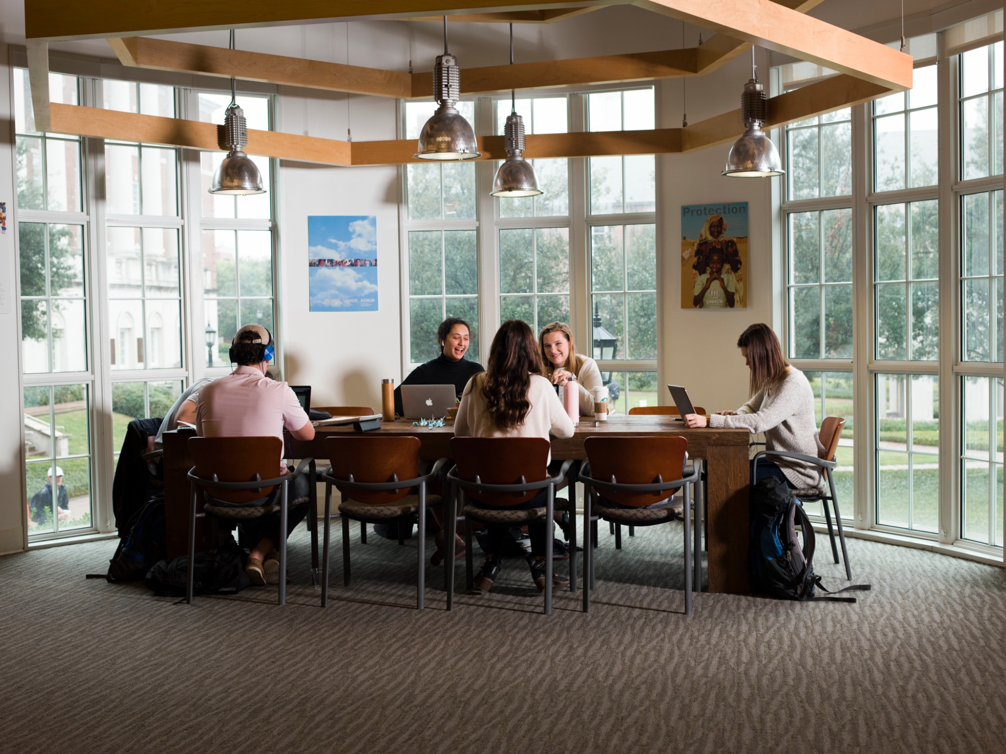 A group of students study together in a bright, modern study space with large windows overlooking a scenic campus. They are engaged in discussions, using laptops and notebooks. The room features wooden ceiling beams, pendant lighting, and wall art. Backpacks are placed around the table.