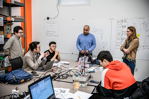 A group of students sits around a table with laptops, collaborating on a project in a classroom setting.